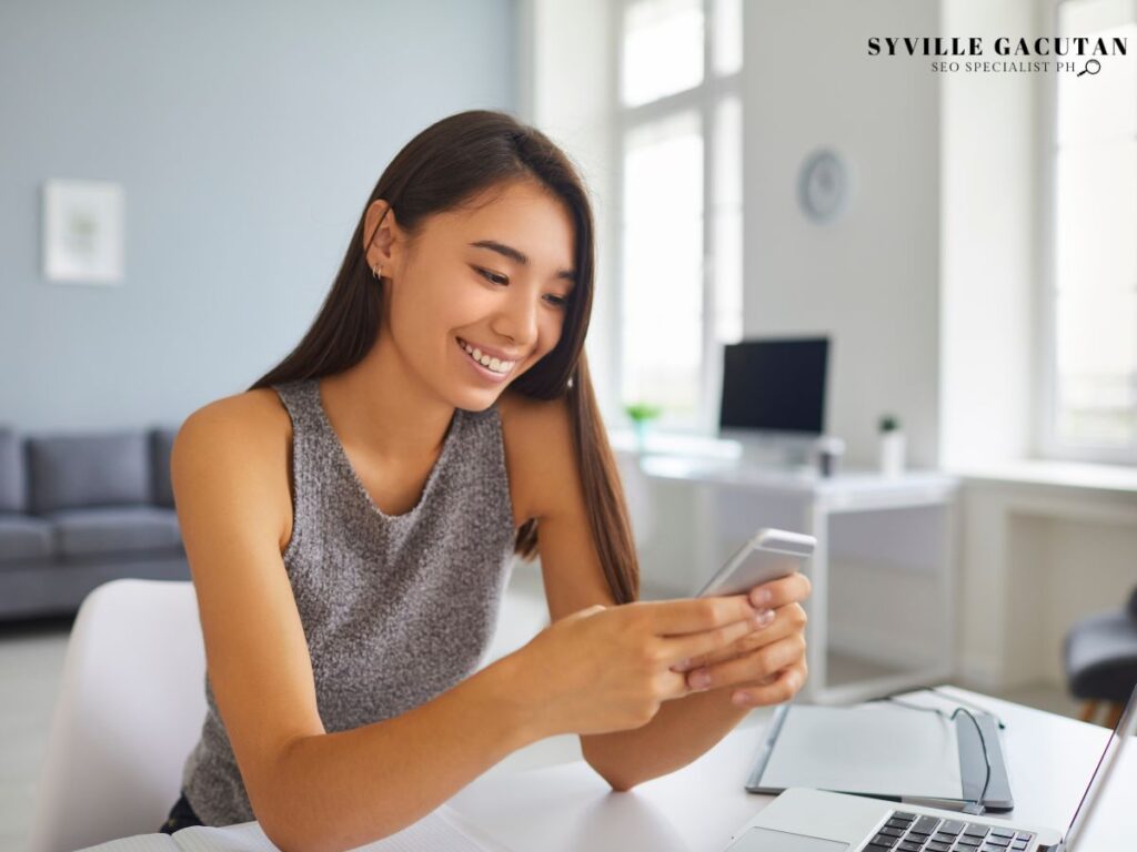 A woman smiling while using her smartphone at a desk.

