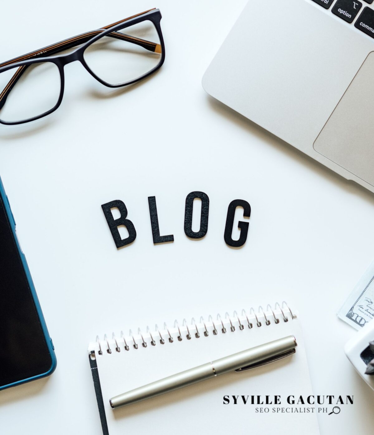 The word "BLOG" surrounded by glasses, a pen, and a laptop on a desk.