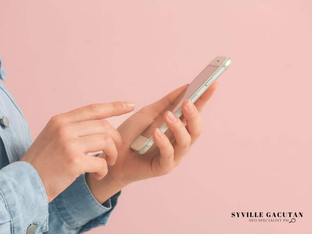 A woman’s hand using a smartphone against a soft pink background.