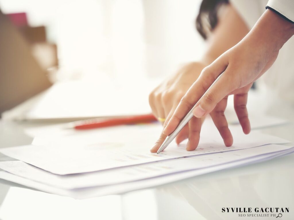 A hand holding a pen pointing at the paper on the desk.