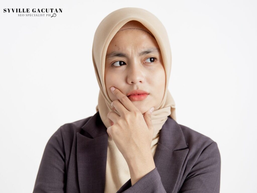 Woman in a beige hijab, thinking with hand on chin, looking thoughtful.