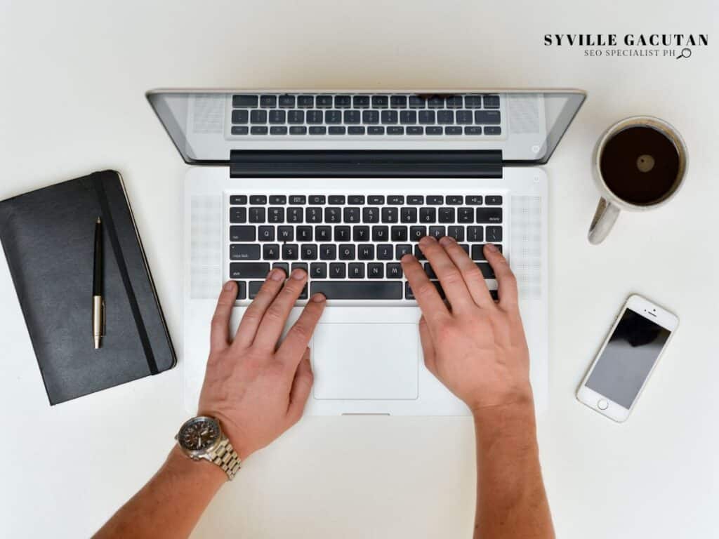 Hands typing on a laptop, with a notebook, pen, phone, and coffee beside it.