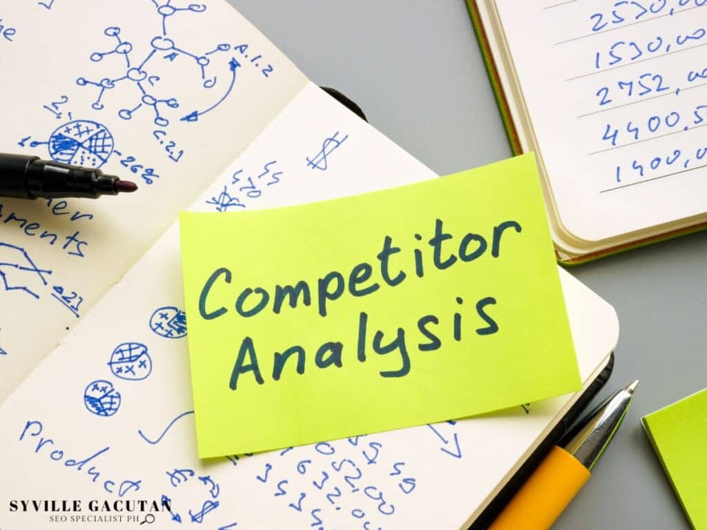 Sticky note reading "Competitor Analysis" on top of notebooks with diagrams.