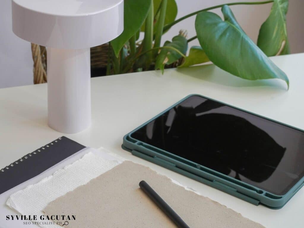 Minimal desk setup with a tablet, notebook, lamp, and plant.