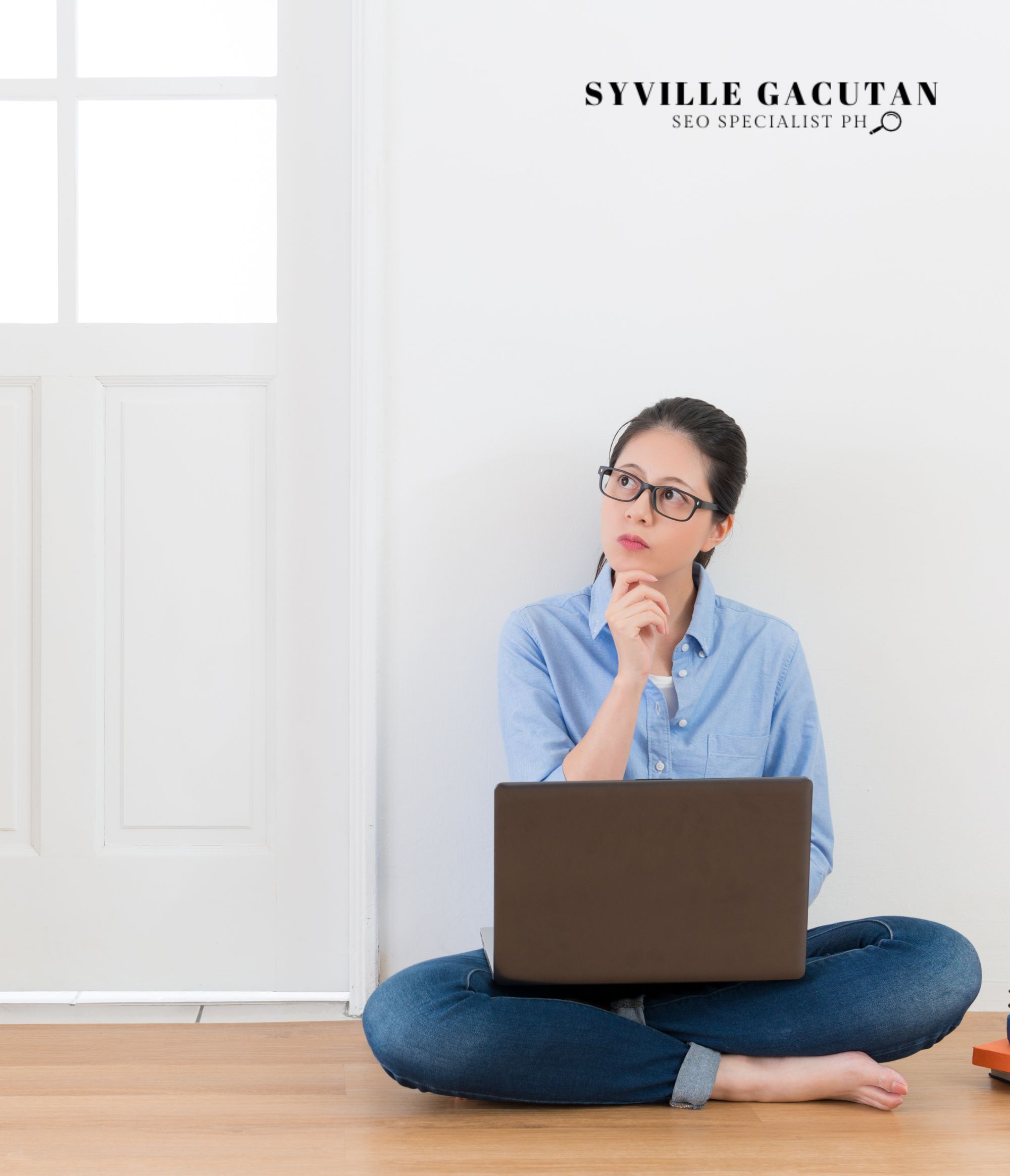 A woman in a blue shirt and glasses sits cross-legged with a laptop, in a minimalist white room.