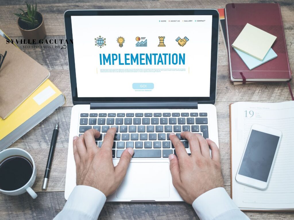 Hands typing 'Implementation' on a laptop with organized desk items.