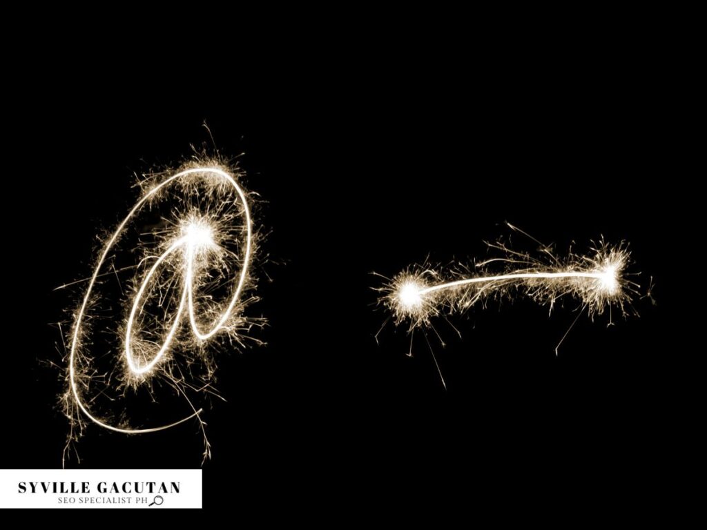 Light trails forming "@" and " - " against a dark background, creating a sparkler effect.