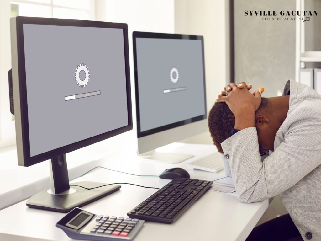 Frustrated worker at desk with loading screens showing gear icons on dual monitors.