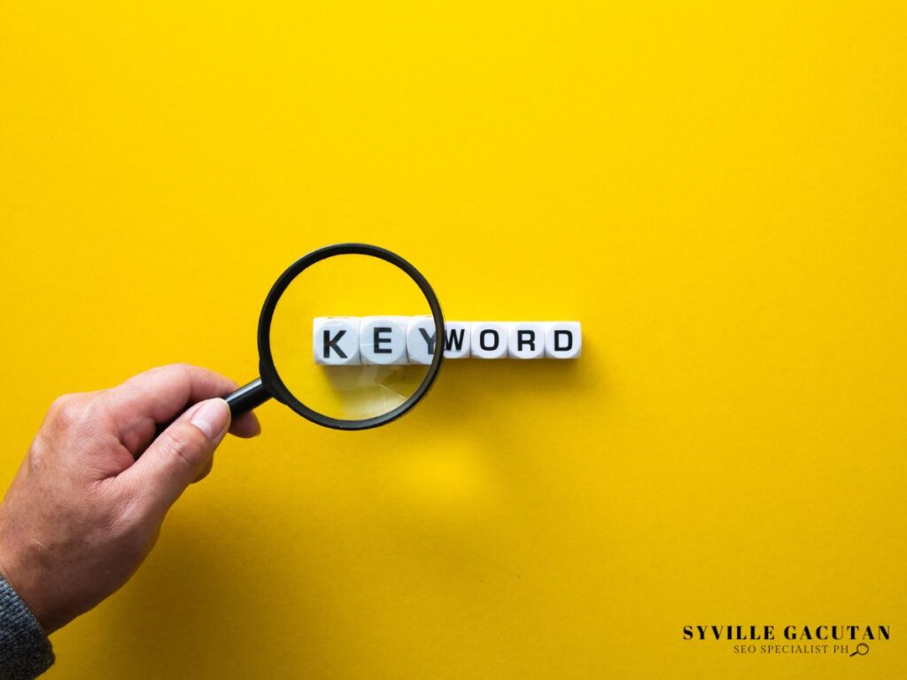 Hand holding magnifying glass over white letter cubes spelling "KEYWORD" on yellow background.