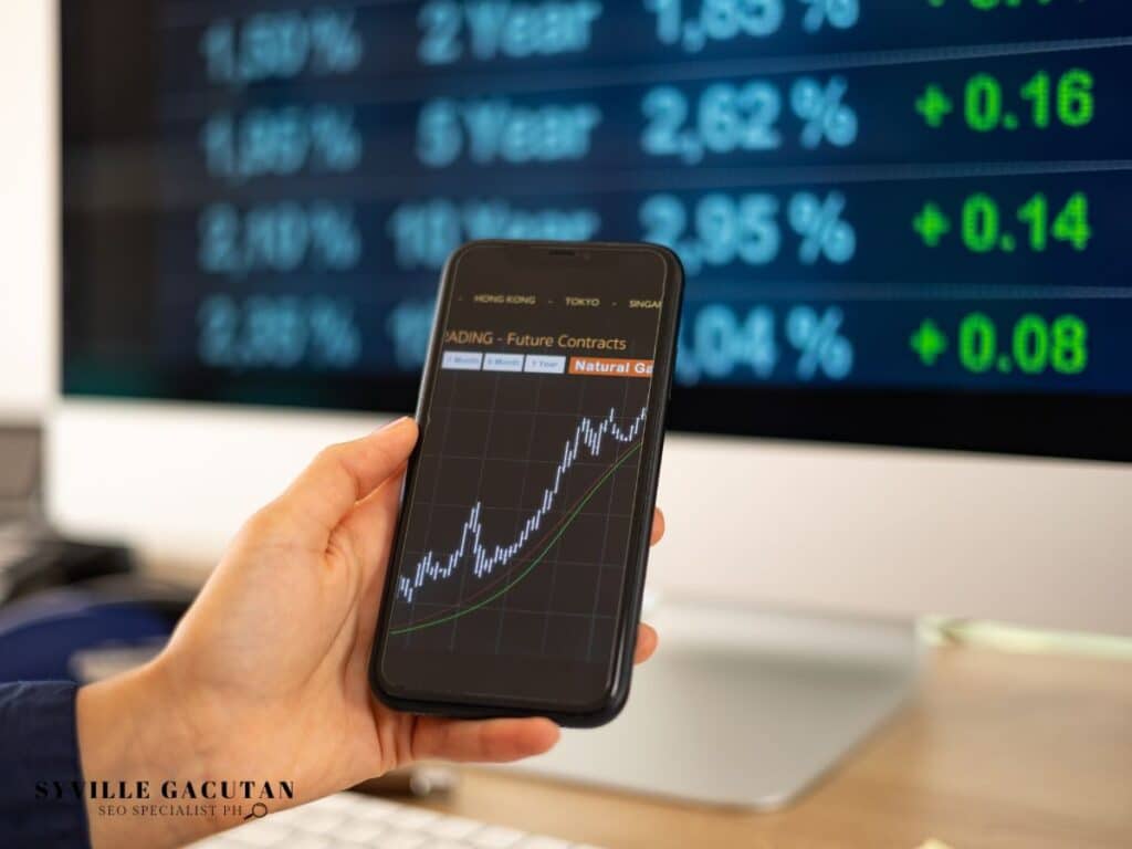 Smartphone displaying a stock chart beside a large monitor.
