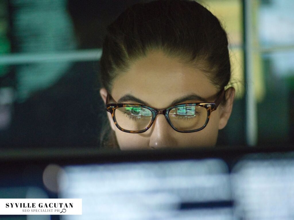Woman focused on screens, wearing glasses with reflections visible.