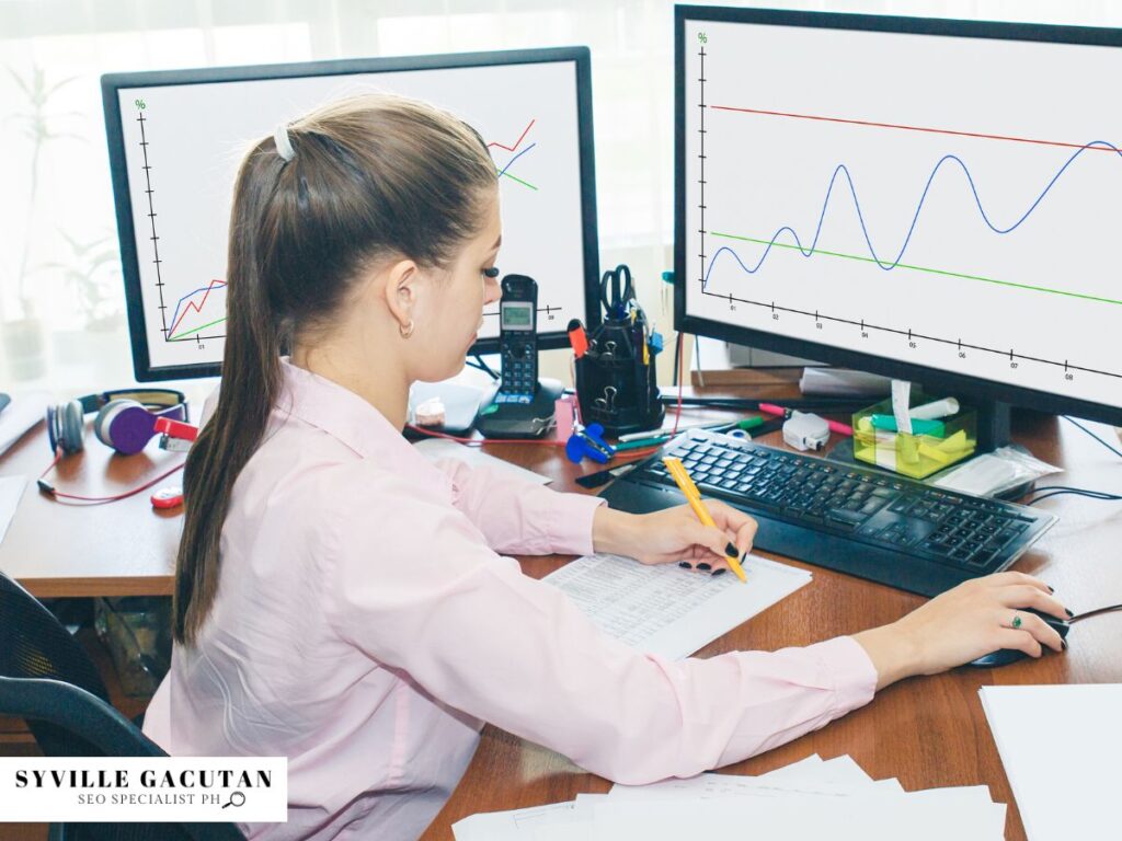 Woman analyzing graphs on dual monitors, taking notes at a desk with office supplies.