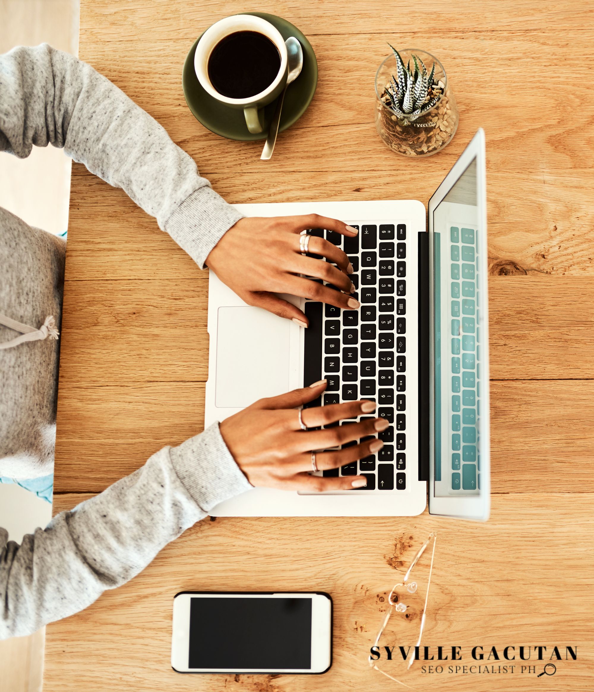Hands typing on a laptop with coffee, a phone, and a small plant on a wooden desk.