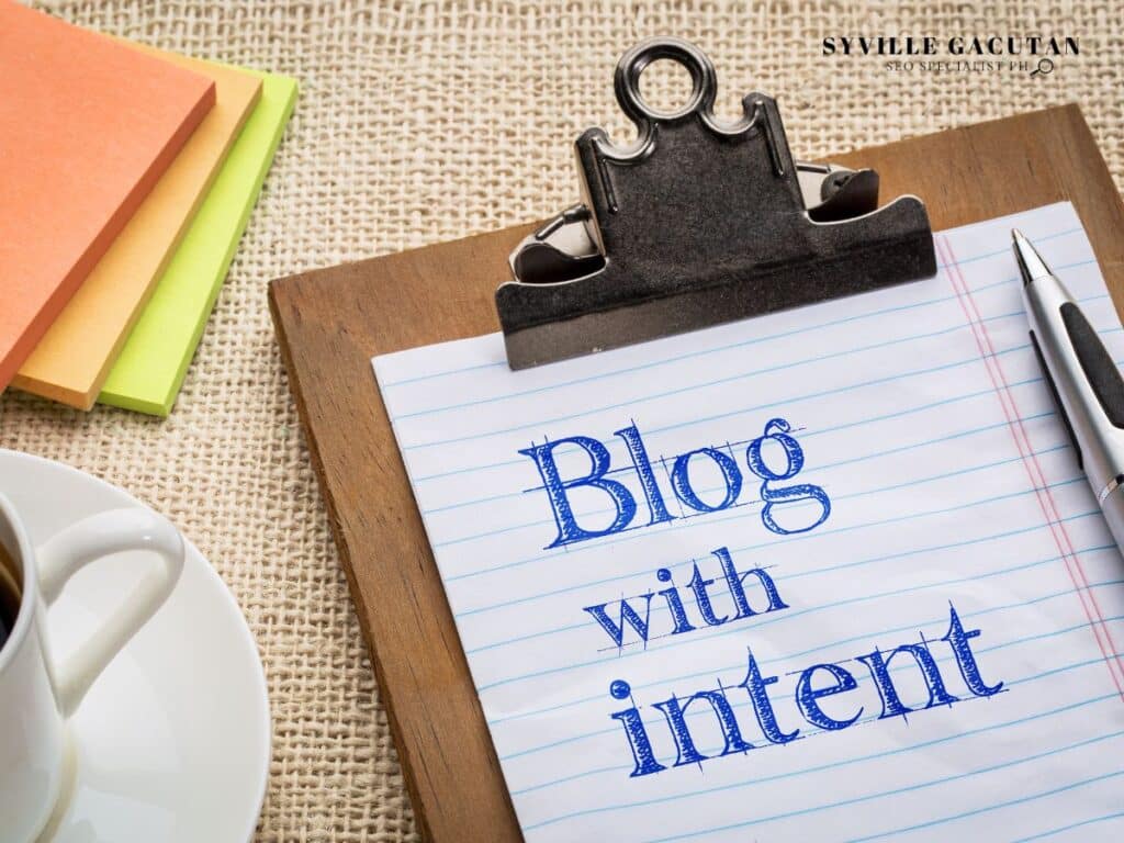 Clipboard with "Blog with intent" text in blue ink, sticky notes and coffee cup on burlap surface.