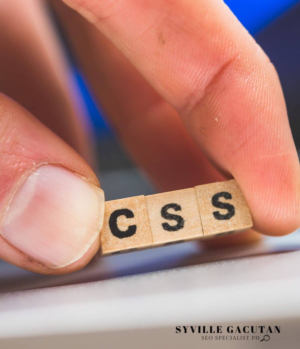 A close-up photo of fingers holding wooden letter tiles spelling "CSS" against a blurred background.