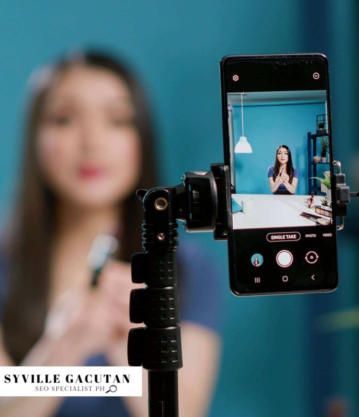 A smartphone on a tripod capturing a photo of a person against a turquoise blue wall.