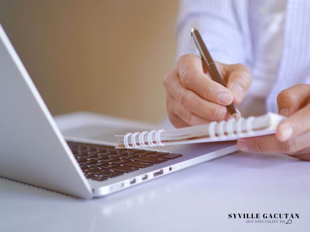 Close-up of hands writing in spiral notebook next to silver laptop keyboard.