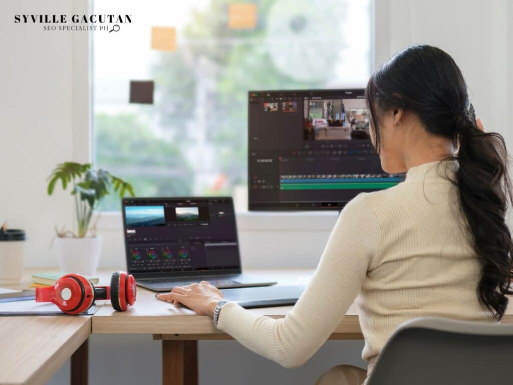 woman working on dual screens with red headphones and cream sweater in bright workspace.