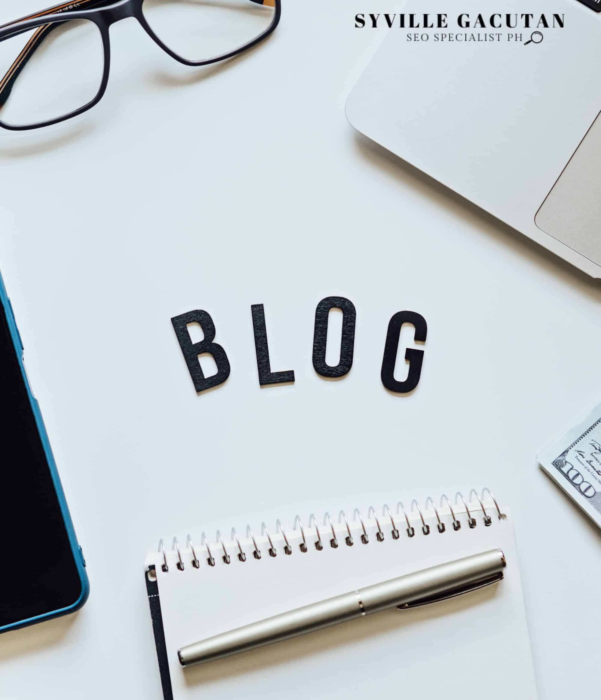 A minimalist desk setup with "BLOG" text, glasses, notepad, pen, and partial view of a phone.