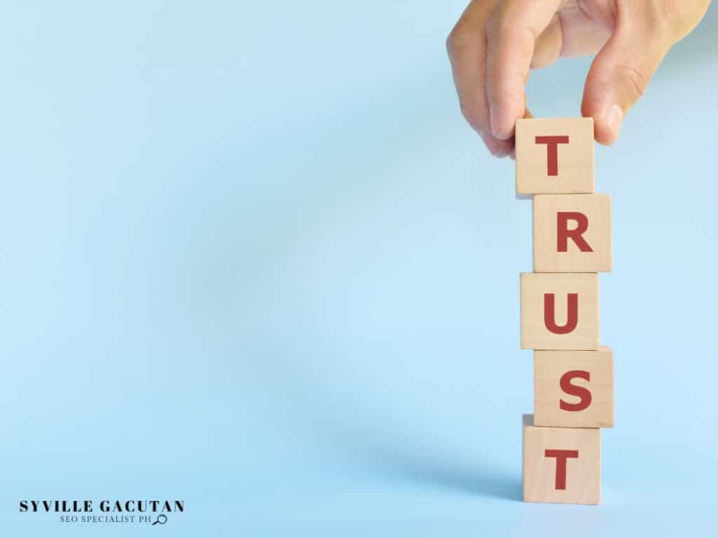 Hand stacking wooden blocks spelling "TRUST" vertically against light blue backdrop.