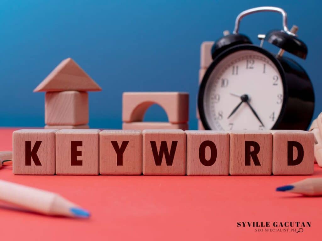 Wooden blocks spelling "KEYWORD" with clock and building blocks on coral pink surface.