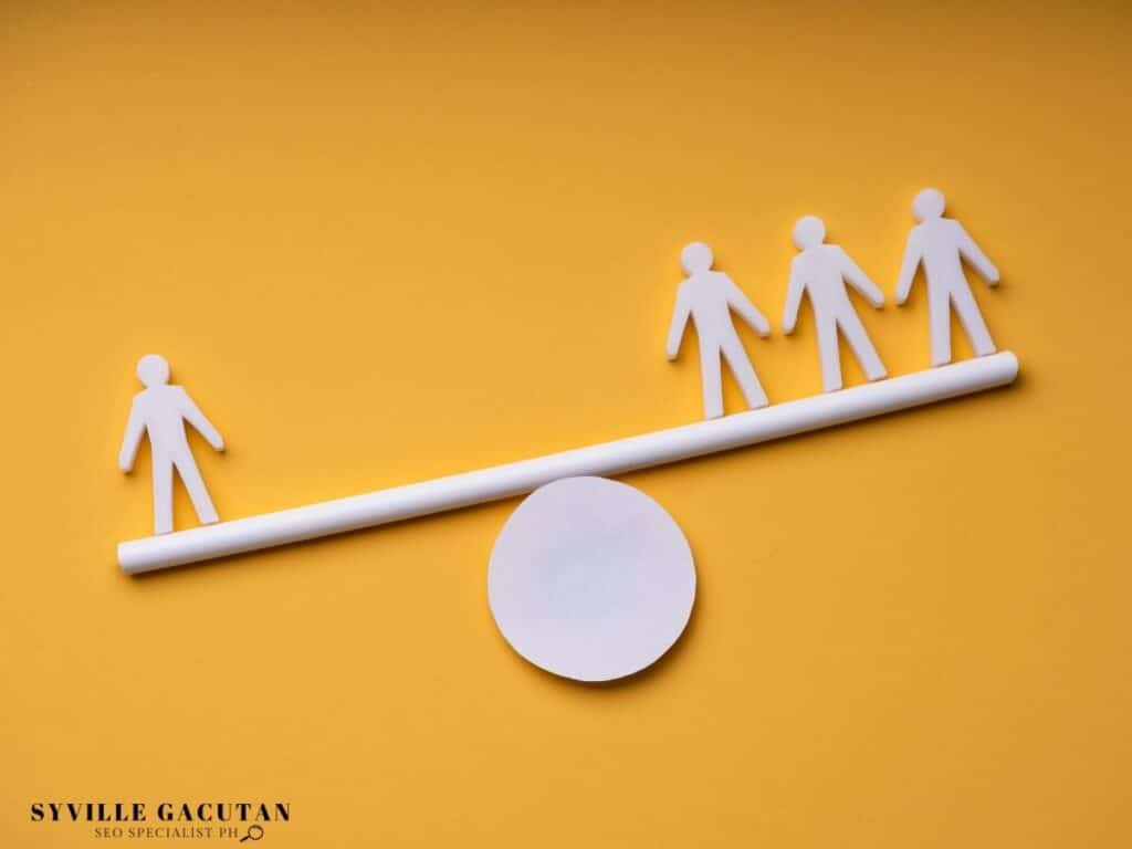 White paper cutout figures balancing on seesaw against yellow background, showing group dynamics.