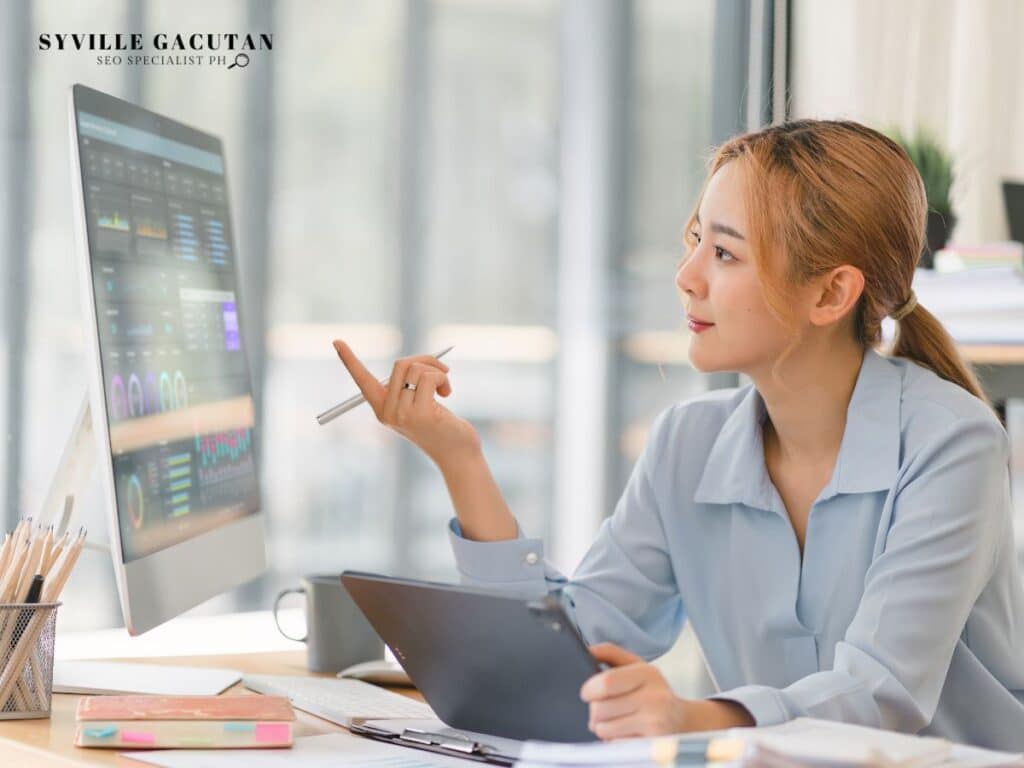 A woman in light blue blouse analyzing data charts on computer monitor in bright office setting.