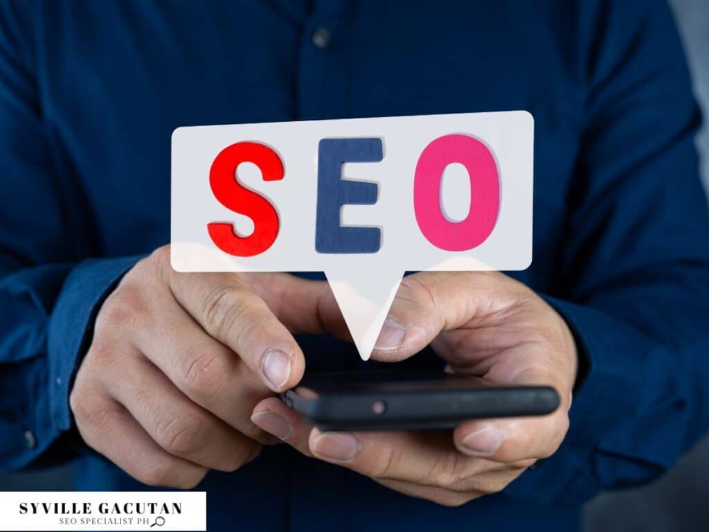 Hands holding phone with white speech bubble displaying "SEO" in red, blue, and pink letters.