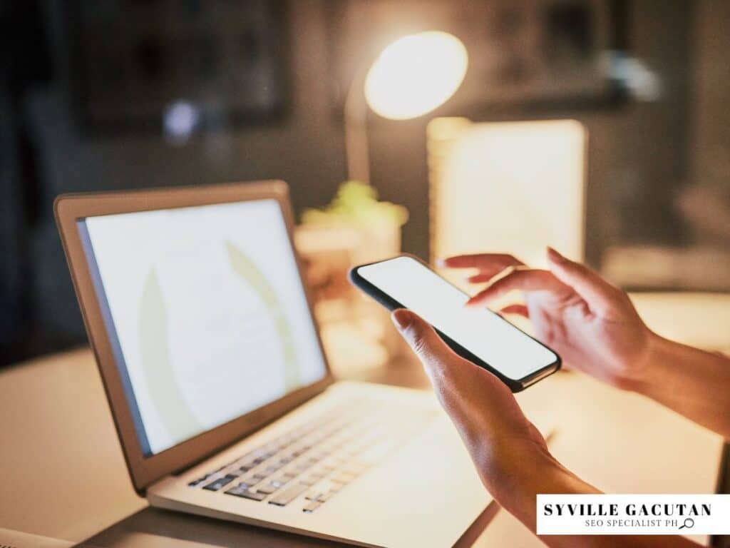 Person using phone and laptop in warm lighting with desk lamp illuminating workspace.