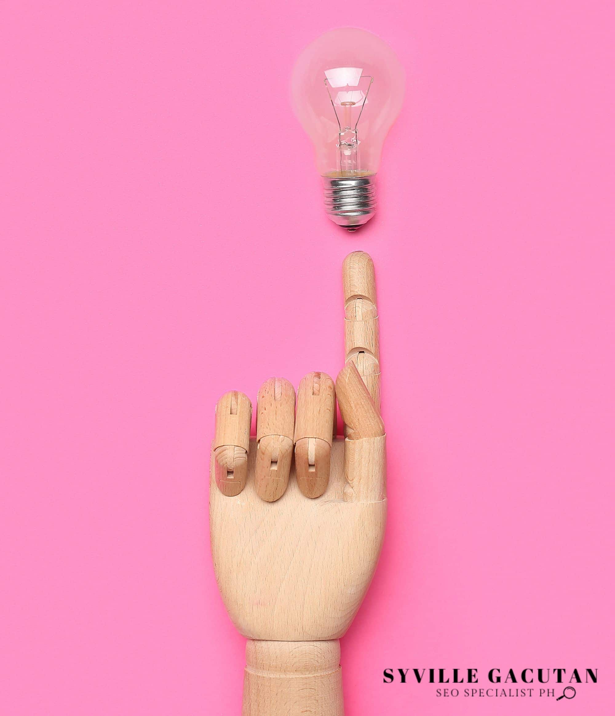 A wooden artist's hand pointing upward at a light bulb against a pink background.