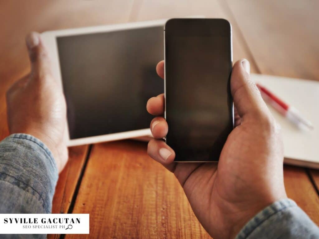 Hands holding smartphone and tablet devices on wooden surface, showing dark screens.