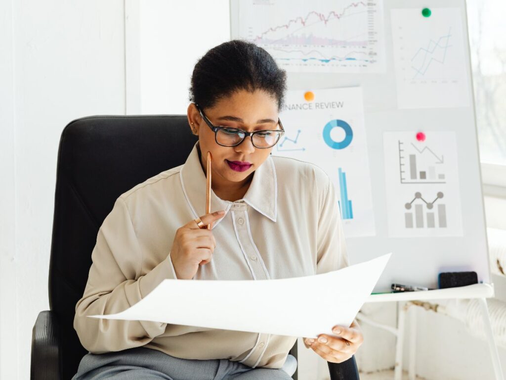 Woman reviewing documents with finance charts behind her.