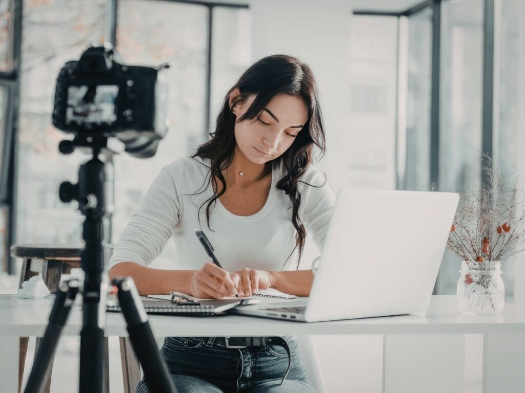 Woman writing content beside a laptop and camera.
