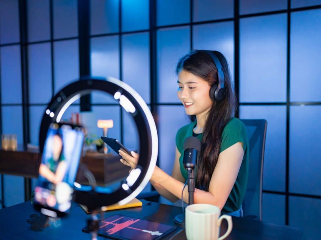 Woman recording content with phone, ring light, headphones, and microphone.