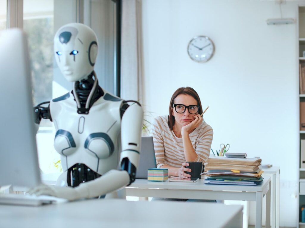 Robot working at a computer while a woman watches thoughtfully.