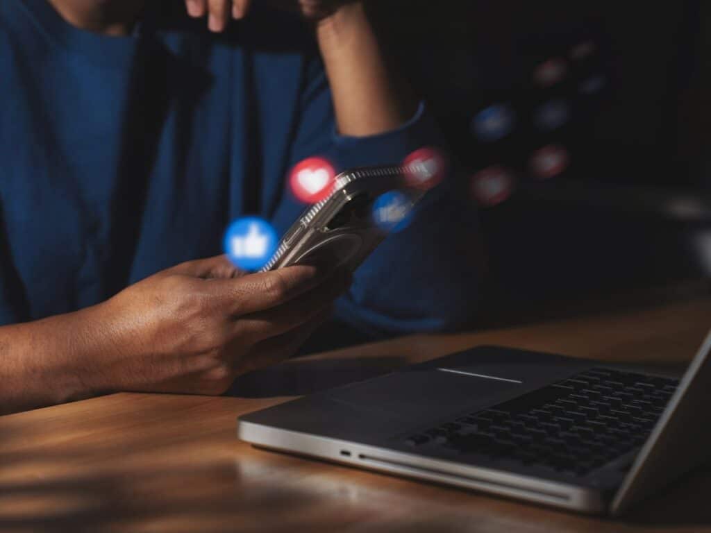 Person using a phone beside a laptop with floating social media icons.