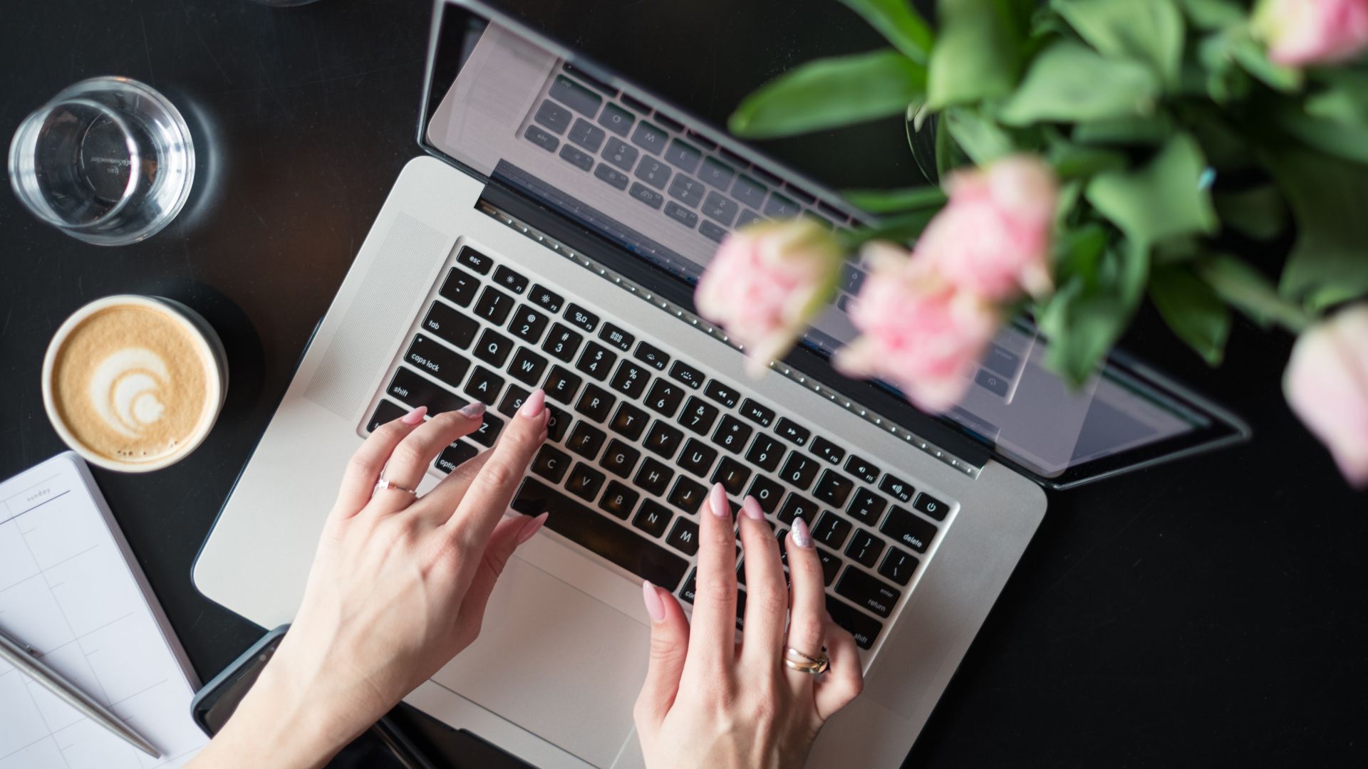 Hands typing on a laptop beside coffee and flowers.