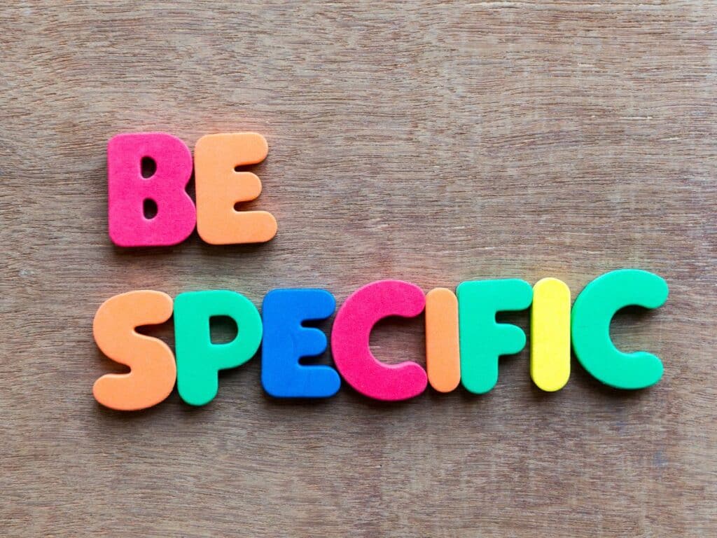 Colorful letters spelling “Be Specific” on wooden background.
