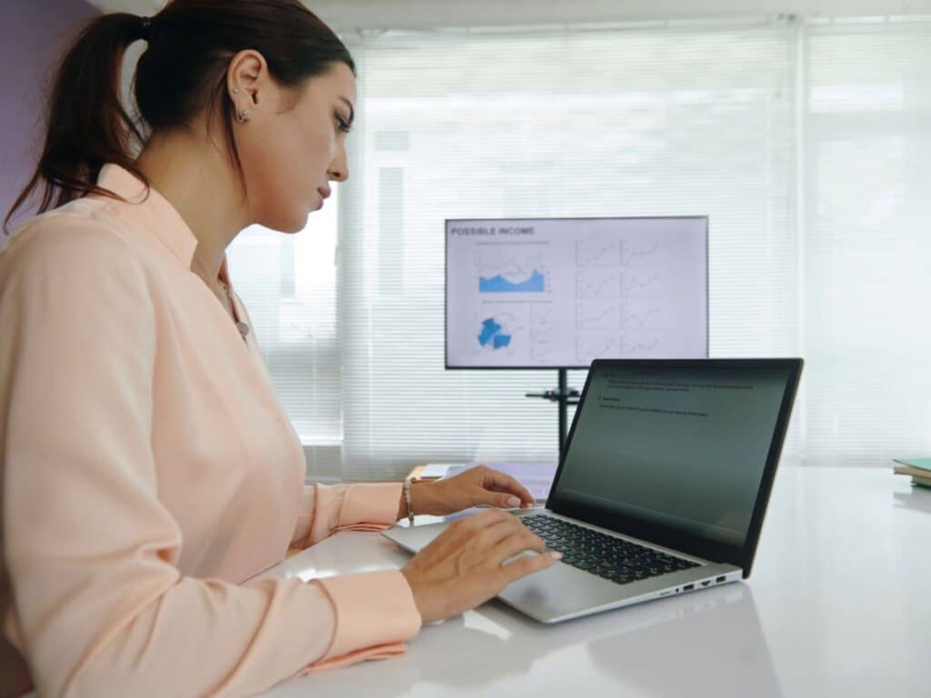 Woman working on a laptop with charts displayed on a screen behind her.