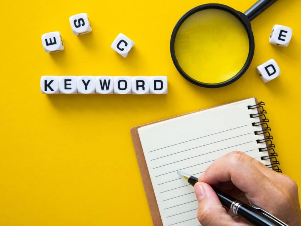 Hand writing in a notebook beside “KEYWORD” blocks and magnifying glass.